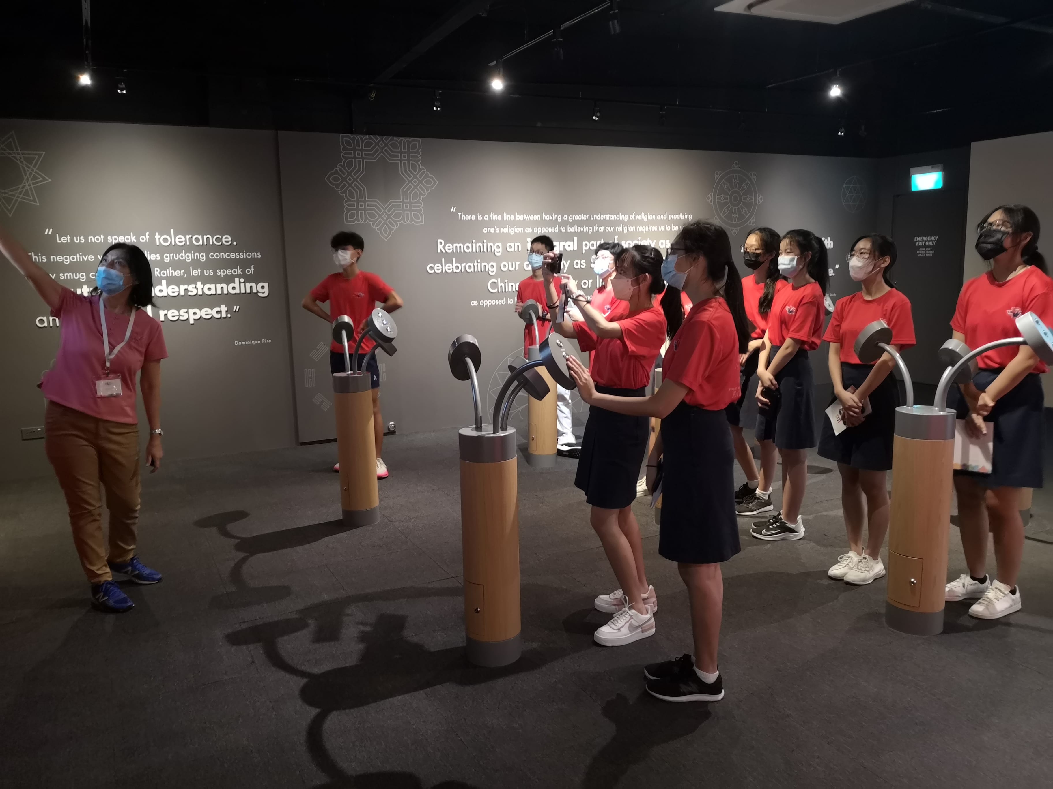 A guide leads masked students in red uniforms through a museum exhibit with interactive pedestal displays and quote-covered walls.